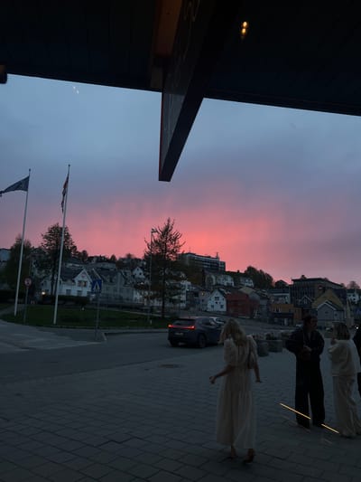 The image shows a street scene in Tromsø, Norway at dusk or dawn with a vibrant pink and purple sky. People are walking on a paved sidewalk near a street with cars. Flags are visible in the foreground near trees and street signs. Buildings are situated on a hillside behind the street. An overhang from a building is in the left corner of the image.