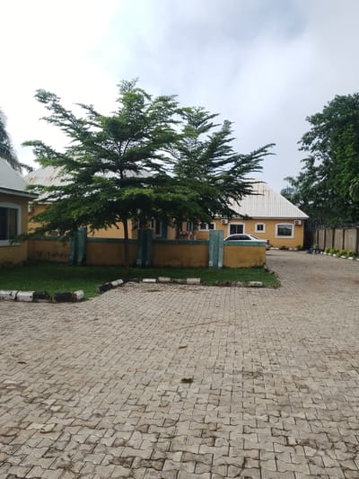 The image depicts a paved courtyard area in front of a single-story, yellow building with a white, gabled roof. A prominent tree with full, green foliage is situated in the foreground, to the left of the building. The paving stones are a light gray, interlocking pattern, showing signs of wear and some staining. A low, yellow wall, topped with decorative teal pillars, separates the courtyard from a grassy area in front of the building. A white car is parked in front of the building. To the right of the building, a concrete fence with barbed wire is visible, with a few people seated on chairs further down the path. The sky is overcast. This scene is likely located in Serti, Nigeria, given the architectural style and the presence of lush vegetation.