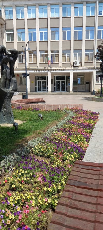 The image shows the exterior of a building, likely a public institution, with a paved area in front featuring a flower bed and a sculpture. The building facade is modern, characterized by multiple rows of identical windows and a light-colored stone or concrete exterior. The inscription above the entrance reads "ОБЩИНА СИЛИСТРА" (Municipality of Silistra), indicating the location as Silistra, Bulgaria.
In the foreground, a circular brick planter contains a vibrant display of colorful flowers, predominantly in shades of purple, yellow, and pink. The flowers are densely planted, creating a lush carpet of blooms. Behind the flower bed, a dark-colored abstract sculpture is partially visible on the left side of the frame. Several dark metal lampposts are strategically placed throughout the scene, and low-lying spotlights are visible amongst the grass and flowers.
The paved area in front of the building shows the entrance with large glass doors, flanked by pillars. Flags are mounted on poles in front of the entrance, and an air conditioning unit is visible mounted on the building wall. A portion of a wooden bench is present in the lower right corner of the image. The overall impression is a well-maintained public space, likely a town hall or administrative building, in the city of Silistra.