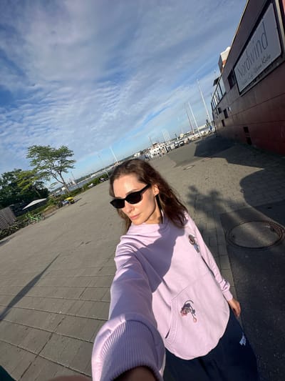A woman with brown hair and sunglasses takes a selfie outside on a sunny day. The location is Kiel, Germany, based on the visible “Nordwind” restaurant sign. In the background are boats docked at a marina and a partly cloudy sky. The woman wears a light purple sweatshirt and dark pants. The ground is paved with grey bricks. A manhole cover is visible.