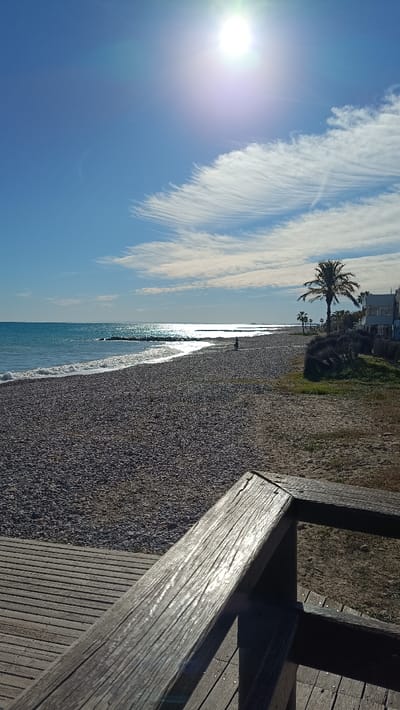 The image depicts a sunlit coastal scene in El Grau De Moncofa, Spain, captured from an elevated perspective overlooking a pebble beach. In the foreground, the top of a weathered wooden railing, part of a boardwalk or viewing platform, extends diagonally from the lower left towards the lower right, partially obscuring a horizontal wooden plank walkway to its left. The sun creates a strong glint on the railing's surface.
The midground is dominated by a wide, light-grey pebble beach that curves gently to the right, meeting the turquoise Mediterranean Sea. Small waves with white foam lines break gently on the shore. A pronounced specular reflection of the sun stretches across the water, creating a shimmering path extending from the horizon towards the beach in the mid-left. In the distant mid-right, a solitary, dark, human-like silhouette is visible standing on the pebble beach near the water's edge. Further out in the water, dark, low-lying structures, possibly breakwaters, are visible.
Along the right side of the beach, a narrow strip of sparse, dry vegetation and grass separates the sand from several white and light-colored coastal buildings with balconies, characteristic of residences. Multiple palm trees are interspersed among these buildings, with one tall, prominent palm tree located in the right midground.
The background consists of a bright blue sky occupying the upper two-thirds of the image. The sun, a brilliant white orb with noticeable lens flare, is positioned in the upper center-right, illuminating the entire scene. Wispy, high-altitude cirrus clouds, exhibiting a feather-like or streaky texture, are scattered across the sky, particularly a distinct formation above the right horizon. The horizon line is visible where the sea meets the distant sky.