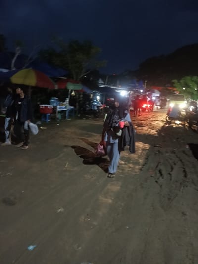The image depicts an outdoor night scene in Kecamatan Pujut, Indonesia, resembling a lively market or gathering area. The sky is dark, characteristic of nighttime, with various artificial lights illuminating the sandy, uneven ground.
In the foreground, a woman, identifiable by her hijab and light-colored attire (possibly a light blue tracksuit), walks from left to right. She holds a red cup, a dark jacket is draped over her arm, and she carries a pink bag. A distinct shadow stretches behind her. To her left, slightly further back, a man in a dark jacket and light pants walks away from the viewer.
The background is filled with informal market stalls, distinguished by colorful umbrellas in shades of red, yellow, blue, and green. These stalls are brightly lit, showing details like coolers and plastic tables. Several people are gathered around these stalls. On the right side of the frame, multiple motorbikes or scooters are present, their headlights and taillights creating bright streaks of light. One vehicle appears to be a three-wheeled transport. Faint outlines of hills are visible against the dark horizon. No visible text is discernible.