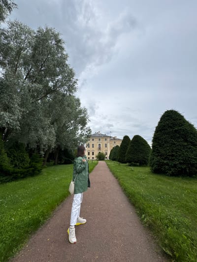 A young woman in a green jacket, white pants, and yellow-accented sneakers stands on a gravel path in a park. She is looking towards a large, pale yellow building with a red flag on its roof, visible in the distance. The path is lined with manicured green grass and several cone-shaped evergreen trees on the right, and a large, leafy tree on the left. The sky is overcast with gray clouds. The building in the background is Constantine Palace, located in Strelna, Russia, on the Gulf of Finland.