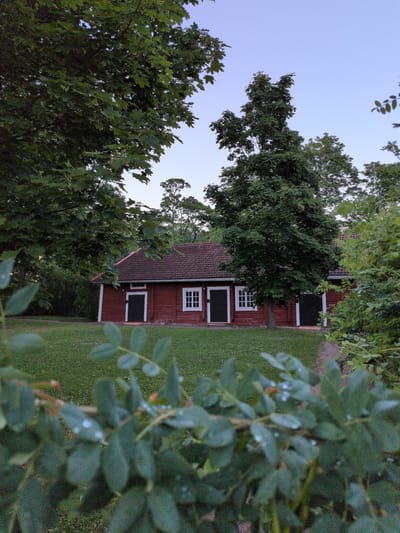The image depicts a small, red wooden building with a dark-tiled roof, situated in a grassy area surrounded by lush green trees. The building has several white-framed windows and dark doors. In the foreground, out-of-focus green leaves with water droplets are visible. The sky above is a clear, pale blue, suggesting it is either early morning or late evening. The overall impression is one of a serene, natural setting. This scene is located at the Finnish Aviation Museum in Vantaa, Finland.