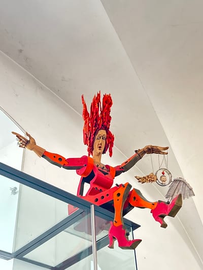 The image captures a peculiar sculpture situated indoors in Heidelberg, Germany. The artwork is suspended from the ceiling and protrudes through a glass barrier. The sculpture is of a figure with flaming red hair, positioned upside down. The body is constructed from red and orange geometric shapes. The figure's arms are articulated, with one hand reaching down and the other raised upward. Above the raised hand is a circular contraption containing a smaller head and adorned with golden wings and what appears to be a fan-like structure. The background consists of a white, angled ceiling and the glass barrier mentioned earlier. The scene conveys a sense of artistic and perhaps surreal display within an architectural space.