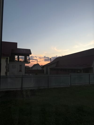 This is a vertically oriented, full shot of a rural landscape at dusk. The sky is a gradient of pale blue to warm orange and pink hues, indicating sunset, with scattered clouds. In the foreground, a dark, grassy lawn is visible on the right side of the image, bordered by a corrugated metal fence. On the left, part of a house with a dark red tiled roof is visible. The house has multiple levels and windows, suggesting a multi-story dwelling or perhaps a building under construction. The overall mood is serene and quiet, characteristic of a rural evening. No people are visible in the frame.