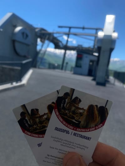 The image depicts two identical business cards held in a person's hand. The cards advertise a restaurant, "Halidzor," which is described as "flying strong for 15 years." The text is in Armenian and English. The background shows a portion of a modern building, possibly a cable car station, with a cloudy sky visible above. The location is likely Shinuhayr, Armenia, given the Halidzor cable car is located there.