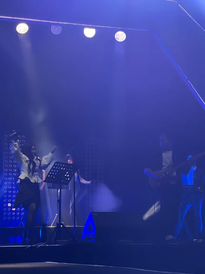 A rotated image of a dimly lit stage in what appears to be an indoor venue. Blue lighting dominates the scene. From left to right: a vertical series of circular stage lights, a person (presumably a singer) behind a music stand, a person on a higher platform with a music stand, and a guitarist on the upper right. The background appears to be an LED light wall. No specific details point to the city of Salembaran, Indonesia.