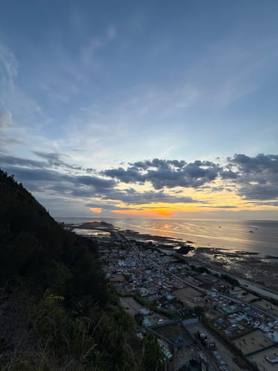 This image captures a coastal settlement at sunset, likely in Lý Sơn Special Administrative Region, Vietnam. The scene is dominated by a panoramic view where the sky meets the sea. The sky is a canvas of dramatic clouds, ranging from dark, heavy formations to wispy, light streaks, all set against a backdrop of fading blue. The setting sun casts a warm, golden glow across the horizon, with vibrant oranges and yellows reflecting on the calm waters of the sea.
Below the horizon, a densely packed settlement of houses is visible, tightly clustered together and stretching along the coastline. The buildings appear to be predominantly small and uniform in structure, with colorful roofs indicating the presence of human habitation. The texture of the land surrounding the settlement suggests a hilly or elevated terrain, with dark green vegetation encroaching on the edges of the frame in the foreground.
The time of day is clearly sunset, indicated by the warm, low-angled light and the color palette of the sky and water. The weather appears to be fair, with scattered clouds adding visual interest to the sky. The location cues, such as the coastal setting and dense settlement, are consistent with descriptions of island communities. The overall mood evoked is one of peaceful twilight, with the natural beauty of the sunset contrasting with the ordered, yet humble, human settlement. There is no visible text in the image.