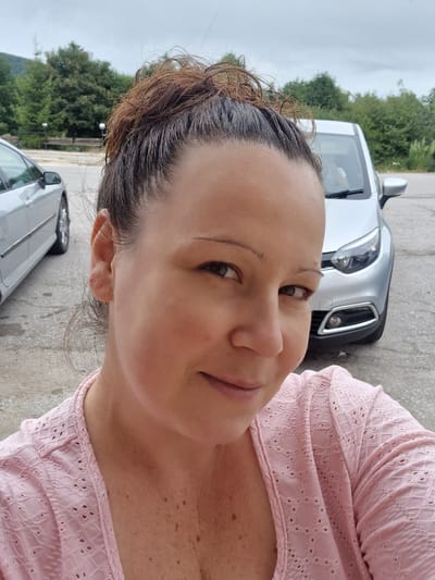 A woman is posing for a selfie outdoors. She has her hair pulled up in a messy bun, and she is wearing a pink top. In the background, there are two cars and some trees. The scene appears to be a parking lot in a natural setting, possibly near a mountainous area, given the presence of trees and a cloudy sky. The location is identified as Shipka, Bulgaria.