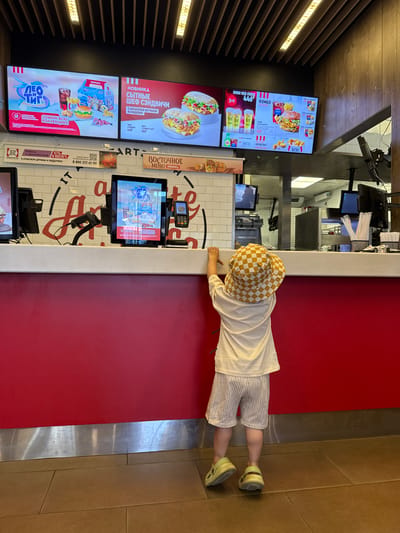 In Заволжье, Russia, a young child wearing a checkered bucket hat and a white t-shirt with striped shorts stands at a red counter in what appears to be a fast-food restaurant. The child is facing away from the camera, looking towards the counter. On the counter, there are two computer monitors displaying menu items and a credit card machine. Behind the counter, a kitchen area is visible with more equipment and monitors. The restaurant interior features a modern design with a wooden ceiling and lighting. The menu boards on the wall display various food and drink options, including sandwiches and combo meals, with Russian text. The overall scene depicts a typical fast-food restaurant setting with a child waiting at the counter.