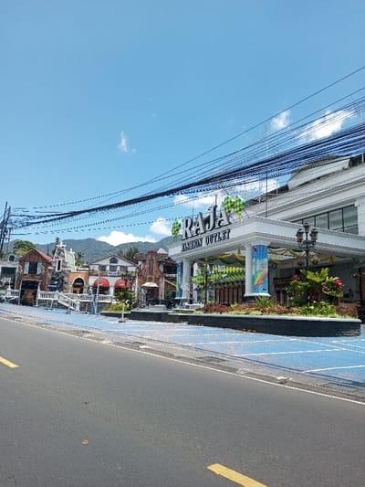 This is a daytime exterior photograph depicting a commercial street in the city of Bagochen Boolingpang Ueling, Bhutan. An asphalt road with a broken yellow center line occupies the lower left foreground. Immediately adjacent to the road, a raised blue-colored paved area with white grid markings extends, serving as a pedestrian zone or parking area. The prominent subject is the "RAJA FASHION OUTLET," a large white building featuring a columned portico entrance. The building's sign, "RAJA FASHION OUTLET," is prominently displayed in metallic letters, surmounted by large green and yellow rhombus-shaped decorations. Green and yellow garlands adorn the entrance portico. In front of the building, a dark planter box contains green and red foliage, and a decorative black street lamp with multiple lights is positioned. To the left of the "RAJA FASHION OUTLET," a series of smaller commercial buildings with diverse architectural styles, some resembling European cottages, line the blue-paved area, with some displaying "RAJA" and "OUTLET" signage. The background features green, tree-covered mountains under a clear blue sky with scattered wispy white clouds. Numerous black utility cables crisscross horizontally across the upper portion of the image. No individuals are visible in the scene.