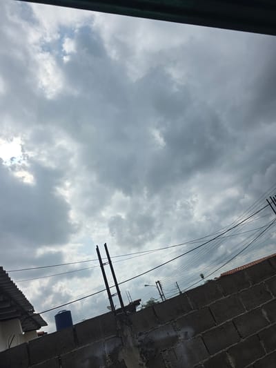 A low-angle shot captures a cloudy sky above Parroquia Ocumare, Venezuela. The foreground is defined by a rough, gray concrete block wall on the right and a dark green vertical structure on the left. Electrical wires and poles extend diagonally across the frame against the backdrop of the clouded sky. Below the wall, parts of a corrugated roof and a blue cylindrical tank are visible. The overall scene conveys a sense of urban infrastructure juxtaposed with a cloudy atmosphere.