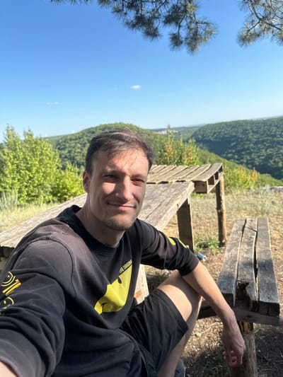 A man in a black long-sleeved shirt with a yellow graphic and black shorts is sitting on a wooden bench. He is smiling and looking at the camera. In the background, there is a wooden picnic table and a lush green landscape of rolling hills and trees under a clear blue sky. The scene appears to be a park or a scenic overlook. The location is identified as Malozalissia, Ukraine.