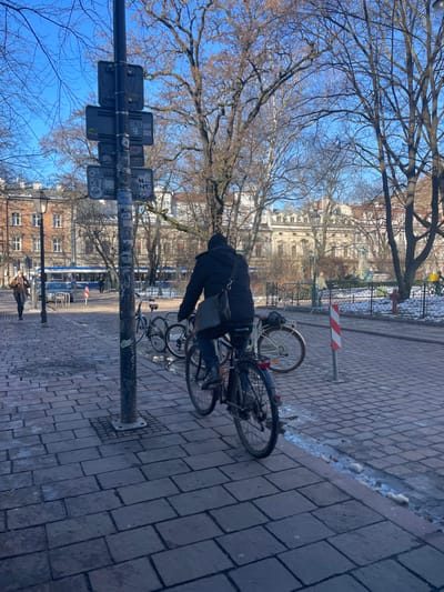 The image depicts an outdoor urban scene in Kraków, Poland, on a bright, sunny day with a clear blue sky. The primary subject is a person, viewed from behind, dressed in a dark hooded jacket and carrying a dark messenger bag over their left shoulder. The person is bent over a bicycle, which is parked alongside several others on a paved sidewalk, suggesting they are either preparing to ride, dismounting, or securing the bike.
To the left of the person, a tall, dark metal pole stands prominently, serving as a signpost or lamppost. It is covered with numerous stickers and has several rectangular signs attached, though no specific text is clearly legible due to the distance and resolution. The sidewalk is constructed from light-colored rectangular paving stones. Along the edge of the curb, patches of snow or ice are visible, indicating that the weather is cold. Further to the right, a red and white striped pole is present, likely functioning as a bike rack or a street barrier.
The background features numerous bare trees, suggesting a late autumn, winter, or early spring season. Beyond the trees, ornate, classical-style buildings with multiple windows and detailed facades can be seen under the bright sky. A faint figure of another person is visible walking further down the street in the distance. The overall scene captures a moment of daily life in a European city, combining urban infrastructure with natural elements.