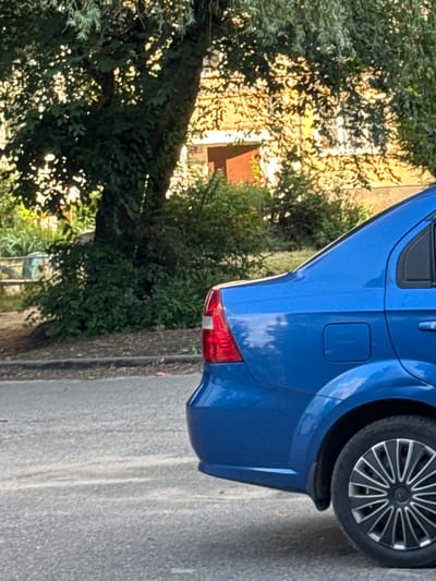 The image shows the rear quarter panel of a blue car parked on a street in Vyazniki, Russia. The car is viewed from a low angle, with its taillight and rear wheel visible. The car is positioned next to a tree with dense foliage, casting dappled shadows on the car's body and the road. In the background, a building with a yellow facade is partially visible through the leaves. The lighting suggests it is daytime, with sunlight filtering through the trees, indicating a possibly sunny day. The scene is static, with no people or apparent activity taking place, suggesting a casual moment of the car being parked. The street appears to be paved, with a concrete curb on the right side of the image. The overall setting is a residential or semi-urban area with greenery. There is no visible text in the image.