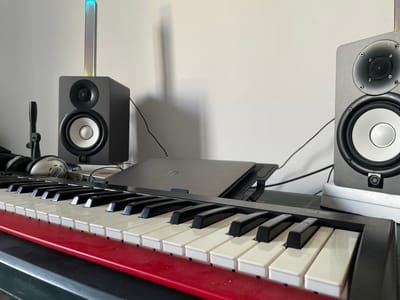 The image depicts a music production setup. In the foreground, a red-trimmed keyboard with black and white keys is positioned on a dark surface. Behind the keyboard, a silver laptop is placed on a stand. To the left of the laptop, a pair of silver headphones rests on the keyboard. Two gray studio monitors are visible on either side of the setup. One monitor is on the left, and the other is on the right. A slim, rectangular light fixture with a gradient of colors (blue, purple, yellow) is placed on top of the left monitor. The background is a plain, light-colored wall, with the shadow of the light fixture cast upon it. The scene is indoors, suggesting a home studio or a dedicated music space. The location is identified as Ustrem, Bulgaria, though no specific city landmarks are visible in the image.