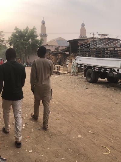 The image depicts an outdoor, unpaved street scene in Gobirawa, Nigeria, under a hazy, light-colored sky, indicative of a dusty atmosphere. In the foreground, two men walk away from the camera. The man on the left wears a dark green t-shirt, light-colored speckled pants, and dark sandals. The man on the right wears a light tan long-sleeved tunic and matching pants with brown open-toed footwear. The ground is reddish-brown dirt, uneven, with visible tire tracks and scattered small debris.

In the midground, to the right of the walking men, a series of rustic, open-front stalls or shops are constructed from wood planks and corrugated metal roofing. These structures contain substantial stacks of cut lumber, wood planks, and wooden crates. Further right, a white truck is parked, featuring a wooden slatted bed frame and dual rear wheels. The letters "NMPC" are visible on its rear mudflap. Transparent panels, consistent with glass or plastic sheets, are stacked within the truck's bed. A person in a light-colored traditional garment is walking from left to right in front of the truck and behind the wood stalls. To the left in the midground, behind a green-leafed tree, a group of individuals are gathered near a building.

The background is dominated by a light tan mosque featuring a multi-faceted roof covered with numerous solar panels. Two identical minarets, each capped with a crescent moon symbol, flank the main mosque structure.