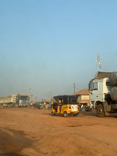 A street scene featuring various vehicles, including a yellow three-wheeled auto rickshaw, large trucks, and smaller cars, are parked along a dirt road. Utility poles line the street, with an antenna towering in the background. The sky is a clear blue, while the ground is dusty. The overall impression is of a busy, possibly roadside, area.