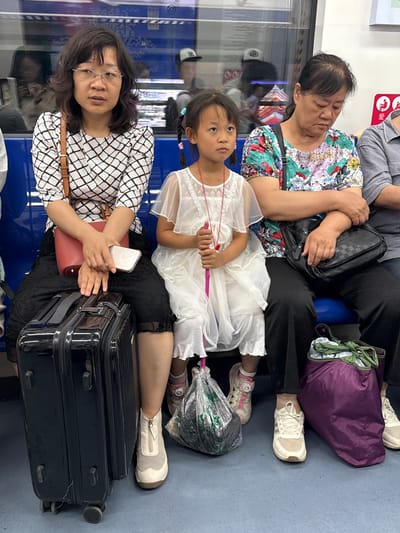 The image shows three people, two adult women and a young girl, seated closely together. The woman on the left is wearing glasses and a black and white patterned shirt. She is holding a silver smartphone in her hands. Next to her, a young girl in a white dress sits between her and another woman. The girl has dark hair pulled back in braids and is holding a pink stick. The woman on the right, partly obscured, wears a colorful floral shirt. Behind them, a purple bag and other luggage are visible. The scene appears to be inside a public transport vehicle, likely a train or bus, indicated by the blue seating and windows. The lighting suggests it is daytime. The people appear to be traveling together.
