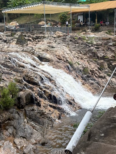 The image depicts a waterfall flowing over a rocky embankment, with a pool of water at the bottom. In the background, there is a structured seating area with people relaxing in a man-made pool under a covered canopy. Further back, a building with a thatched roof serves as a reception or service area, with more people visible on tiered steps. Lush green vegetation is present throughout the scene. The overall setting suggests a natural attraction with developed amenities for visitors. The location is identified as being in Khanh Vinh, Vietnam.