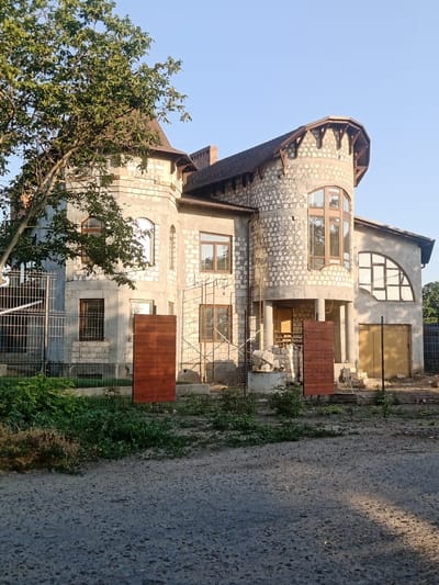 The image depicts a large, unfinished stone house with a brown roof, located in Bender, Moldova. The house features a distinctive round tower with large arched windows and wooden beams supporting the roofline. Adjacent to the tower, a conical turret with a brown roof is visible. The exterior walls are constructed of light-colored stone blocks, and some sections appear to be covered in stucco.
In front of the house, there are several construction materials, including stacks of white bricks and wooden planks. A metal fence encloses part of the property, and a large tree with green foliage stands to the left of the house. The ground in front of the house is covered with gravel. The sky above is clear and blue, indicating daylight. The scene suggests that the house is under construction or undergoing renovations.