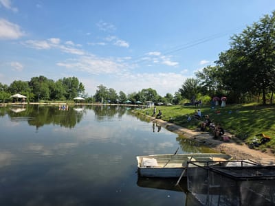 The image shows a serene fishing spot by a lake, possibly at the "Fisherman's Village" in German, Bulgaria, given the context of the query, on a clear, sunny day with scattered white clouds. The foreground features the dark, reflective water of the lake with a small, light-colored boat moored to a wooden platform. A metal cage-like structure is also partially visible on the platform.
The middle ground depicts the lake's shoreline, which is lined with green grass and a gravel path. Several people are seated along the bank, engaged in fishing. They are scattered, some alone and some in small groups, with fishing rods extended towards the water. Umbrellas and tents are set up along the far bank, providing shade. In the background, a line of lush green trees borders the lake, creating a natural backdrop. The sky above is a vibrant blue. The overall scene conveys a peaceful atmosphere of recreational fishing.