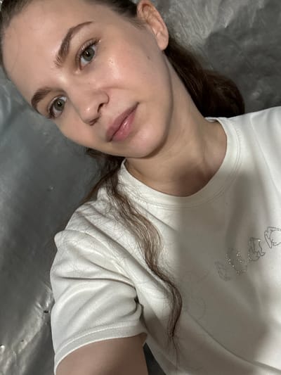 A young woman with light brown hair and green eyes is positioned in the lower right of the frame, looking towards the camera. She is wearing a white t-shirt with a graphic design on the left side of the chest. Her skin appears smooth and slightly luminous. The background is a textured, silvery surface that appears to be a wall or curtain. The overall lighting is soft and diffused, creating a gentle glow on the subject's face. There are no visible indications of the location being in Nizhnyaya Gar', Russia.