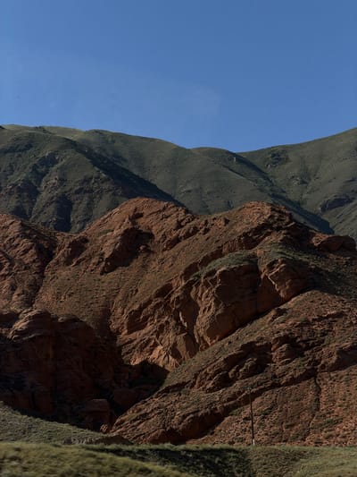 The image displays a mountainous landscape under a clear blue sky. The foreground features a slope covered in sparse green vegetation. Behind this, a prominent ridge of reddish-brown rock dominates the mid-ground, showing rugged textures and geological formations. Further back, a higher mountain slope is covered in dense, dark green vegetation. The scene appears to be a natural, undeveloped area, likely within a rural or wilderness setting. There are no visible people, structures, or vehicles in the image. The overall impression is one of a vast, arid, and natural environment.