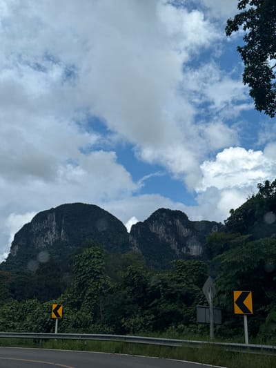 A photograph captures a dramatic landscape with two prominent, dark, rocky mountains dominating the background, their surfaces textured with vertical striations and covered in dense green vegetation. A partly cloudy sky with patches of blue fills the upper portion of the frame. In the mid-ground, lush green foliage and trees form a thick forest that extends from the base of the mountains to the foreground. A metal guardrail runs horizontally across the lower part of the image, separating the roadside from the embankment. To the left of the guardrail, a yellow and black road sign with a left-pointing arrow is visible. Further to the right, another similar sign is present, also indicating a leftward turn. A paved road with a yellow dividing line is in the immediate foreground, suggesting a scenic route. The image appears to be taken from a vehicle, as indicated by the slight blur and the perspective. The scene is situated in Ban Pak Ra, Thailand, a location known for its karst topography and natural beauty.