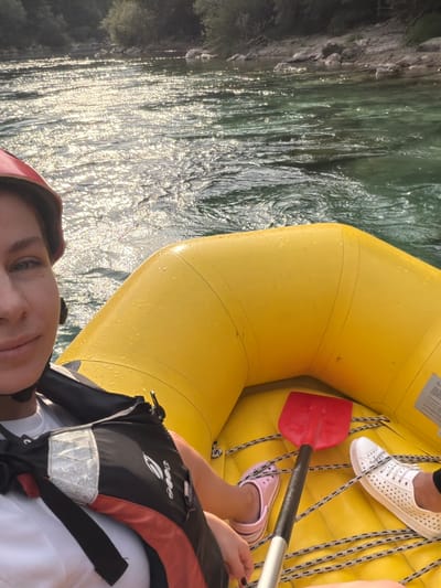 In Polje Bijela, Bosnia and Herzegovina, a person with a red helmet and a life vest is in a yellow raft on a body of water. The water is clear and green, reflecting sunlight, with trees and rocks visible on the shore. A red paddle and white shoes are inside the raft. The person is looking at the camera, and the image is taken from their perspective.