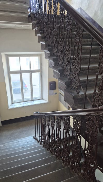 The image shows a view looking down a concrete staircase with ornate, dark metal railings. The staircase appears to be in an indoor setting, likely a building's interior, such as an apartment building or a public space. The railings are highly decorative, featuring intricate scrollwork and repeating patterns, suggesting an older architectural style.

The staircase has multiple flights, descending into the lower part of the frame. The concrete steps show signs of wear, indicating frequent use. To the left of the staircase, there is a cream-colored wall and a portion of a wooden door or paneling.

Below the staircase, a large window with multiple panes is visible. Through the window, it is possible to see some objects, including what appears to be a red and blue toy or object, and possibly a person or their reflection. The window looks out onto an outdoor area, but the details are obscured by the glass and lighting conditions.

On the wall near the window, there is a small, dark square object, possibly an intercom or electrical box, and some papers attached to the wall. The walls themselves are painted a pale yellow. The overall lighting suggests it might be daytime, with natural light coming through the window.

There is no visible text in the image that can be clearly deciphered. The scene evokes a sense of everyday life within an older building, with a focus on the architectural details of the staircase. The location context of Daugavpils, Latvia, could imply a setting in Eastern Europe, potentially contributing to the building's architectural style.