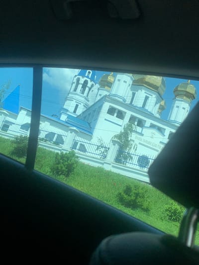 The image shows a white building with blue accents, framed by a car window. The building, located at latitude 49.991, longitude 36.054 in Solonytsivka, Ukraine, appears to be a church or religious institution, featuring multiple gold domes and a sign that reads "Церква Зачаття Святого Івана Предтечі," which translates to "Church of the Conception of St. John the Baptist." The building is situated amidst lush green foliage. The camera is positioned inside a vehicle, looking out the window. The image captures the exterior of the building and some surrounding vegetation.