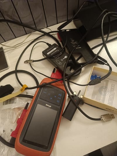 A jumbled collection of electronic diagnostic tools and cables is spread across a white, textured surface, likely a desk or countertop. The central object is an orange and black handheld diagnostic scanner, identified by the "XTOOL" logo. Various black cables snake across the scene, some coiled and others connecting to different devices. A black rectangular adapter with a sticker reading "S/N www.xtooltech.com" is visible, as is a black, textured grip tool. A small cardboard box contains a blue connector and a certification document. The background features a dark, possibly black, ribbed surface. The overall impression is one of a workspace cluttered with automotive diagnostic equipment. The scene is likely indoors, given the artificial lighting and the nature of the objects. There are no visible people or indications of a specific location in Huntingdonshire, United Kingdom.