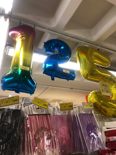 The image, captured in Marousi, Greece, depicts an indoor retail setting, likely a party supply store or a section of a larger store. The photograph is rotated 90 degrees clockwise, causing items that would typically hang vertically to appear horizontally.
The main subjects are several large, inflatable foil balloons and stacks of packaged party decorations. In the upper part of the frame, two golden foil balloons are visible. One, resembling the letter 'M' or a number, has a price tag displaying "ΔΕΙΓΜΑ 10" (Sample 10) and "E 1.99 A" (1.99 Euros). Another gold balloon is partially visible.
Below the golden balloons, a blue foil balloon, shaped like the number '2' or the letter 'S', is prominently displayed. It has a smaller, less clear price tag, possibly reading "75 A". Further down, a vibrant rainbow-colored foil balloon, resembling the letter 'L' or 'I', is visible. A tag on this balloon reads "ΔΕΙΓΜΑ 3" (Sample 3) and "E 1,49 A" (1.49 Euros).
To the right side of the frame, several stacks of packaged party decorations are displayed on shelves. These packages contain what appear to be decorative fringes or streamers in various colors, including purple, pink, red, and black. Each package has a circular cut-out window, revealing the texture of the contents. Multiple price tags are attached to these packages, consistently showing "E 1,49 A" (1.49 Euros). One tag also includes Greek text: "ΕΞΩΦΥΛΛΟ ΚΟΛΛΗΣΗ 20Χ30 ΠΕΤ. ΓΙΑ...", which translates to "Cover sticker 20x30 PET for...". The background shows a light-colored ceiling with recessed lighting fixtures, indicating an indoor, brightly lit environment. No people are visible.