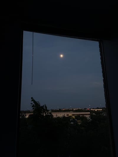 The image shows a view from a window in the city of Austin, Texas. The sky is dark, illuminated by a bright moon. A structure is visible through the window, as are other buildings with illuminated windows and street lights in the distance. Trees are visible at the side of the window.