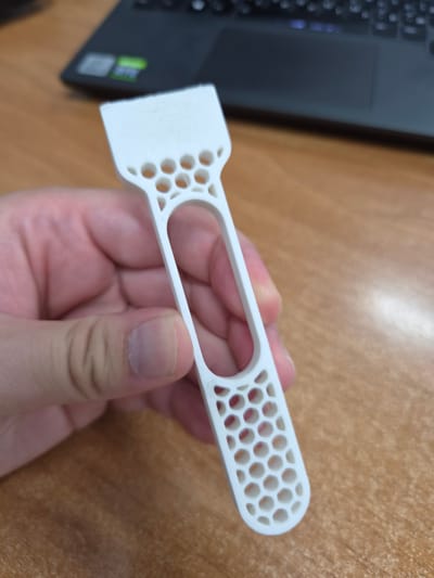 A white, 3D-printed object with a hexagonal cutout pattern is held in a person's hand. The object has a flat, wide end and a narrower, elongated body with a large oval cutout. The person's fingers are visible, gently gripping the object. In the background, a laptop with a black keyboard is resting on a wooden desk. The scene appears to be indoors, with the wooden surface suggesting a desk or table. The image is a close-up shot, focusing on the object and the hand holding it. There is no indication of the location within the city of Kamianske, Ukraine, as the scene is generic and lacks any identifying landmarks or context.