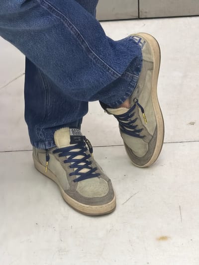 A person wearing blue jeans is standing on a white tiled surface. The person is wearing a pair of Golden Goose Deluxe Brand sneakers with blue laces. The scene is indoors, the sneakers and jeans are in focus, and the background is a plain white tiled floor. It is not possible to determine the location in Santa Clara Ocoyucan, Mexico, from the provided image.