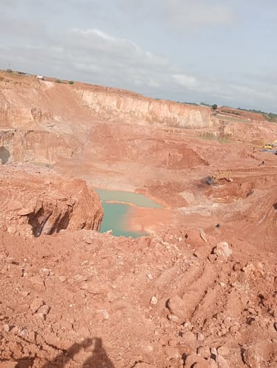 The image shows an active quarry site in Kwang, Nigeria. The terrain is characterized by large, exposed red clay formations with steep, uneven surfaces. Excavation activities are evident, with heavy machinery, including yellow excavators, present and operational within the quarry. Puddles of turquoise water have formed in excavated areas, suggesting the presence of groundwater or collected rainwater. Several figures, appearing to be workers, are visible near the machinery and in the quarry. The sky is overcast with clouds. The foreground is filled with loose reddish soil and rocks, indicating recent excavation or erosion. In the distance, parts of the quarry extend upwards with more layered formations, and some structures and a truck are visible at the upper edge of the quarry.