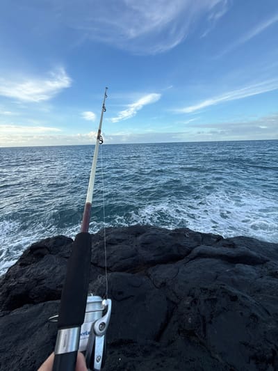 The image displays a first-person perspective of a fishing activity from a dark, rugged, rocky coastline in Hitiaʻa ʻo te Rā, France. A human hand is visible in the bottom left, gripping the black foam handle of a fishing rod. A silver spinning reel is attached to the rod, with fishing line extending up the rod's length. The two-tone fishing rod, primarily light-colored with a brown segment, extends diagonally upwards towards the open ocean. The immediate foreground consists of wet, dark, irregular rock formations. Beyond the rocks, the ocean stretches to the horizon, exhibiting choppy, dark blue water with white foam from breaking waves. The upper half of the image features a bright blue sky with scattered, wispy white cirrus clouds. No other individuals or vessels are visible in the scene.