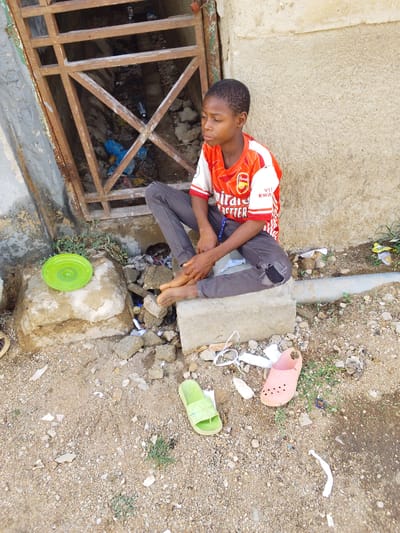 An adolescent boy in an Arsenal soccer jersey sits cross-legged on a concrete block outdoors in Bauchi, Nigeria. He is wearing gray pants with a hole in the knee and is barefoot. Adjacent to him is a metal grate covering a trash-filled opening, a green plastic plate on a rock, and two discarded sandals. The ground is dirt and gravel.