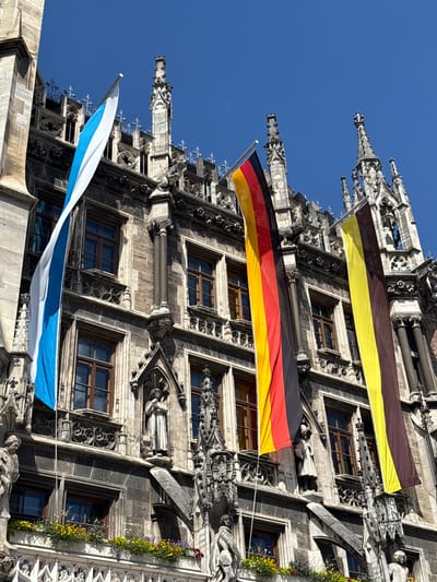 The image shows the Neues Rathaus (New Town Hall) in Marienplatz, Munich, Germany, on a clear day. Three large flags are displayed on the facade of the building. From left to right, they are the flag of Bavaria (blue and white), the flag of Germany (black, red, and gold), and a flag with horizontal stripes of yellow and brown, possibly representing the state of Bavaria or a historical flag. The architectural style of the building is Gothic Revival, characterized by its intricate stonework, spires, and statues. Multiple windows with wooden frames are visible, and a row of flower boxes with yellow and purple flowers adorns a lower balcony. Several stone statues are embedded in the facade. The sky is a bright, cloudless blue.