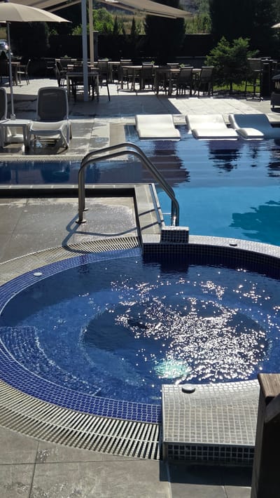 An outdoor swimming pool and hot tub area are shown, likely in Bania, Bulgaria, judging by the context. In the foreground, a circular hot tub with blue tiled edges and a water feature is visible, with sunlight glinting off the water's surface. To the left and behind the hot tub, a section of a larger swimming pool can be seen, featuring submerged lounge chairs. A stainless steel ladder provides access to the pool. Beyond the pool area, lounge chairs and tables with umbrellas are set up on a tiled patio, with trees and buildings in the background. The scene is bathed in sunlight, indicating a clear day. There are no people visible in the image.