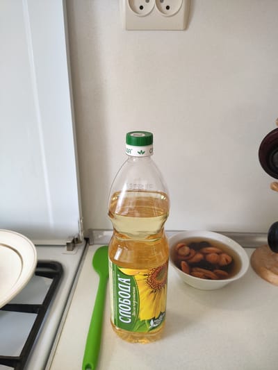 This is a close-up, eye-level shot of a kitchen counter in Navapolatsk, Belarus. The focal point is a clear plastic bottle of sunflower oil, labeled "СЛОБОДА," with a green cap. The oil is golden yellow and fills about two-thirds of the bottle. Next to the bottle is a white bowl filled with dried fruit, possibly apricots and prunes, in a light liquid. A bright green silicone spatula rests on the counter to the left of the oil bottle. In the background, a white wall is visible with a beige electrical outlet and a portion of a white appliance, possibly a refrigerator, on the left. The counter surface is light-colored and appears to be laminate. The scene is well-lit, suggesting indoor lighting. There are no people or animals present.