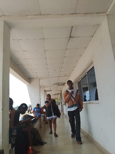 Here is a description of the image:

Location: Covered outdoor corridor in Umueshi, Nigeria.

Environment: Ceiling tiles are visible, with white-painted walls and pillars. Windows are present on the right-hand side of the corridor.

People: Several individuals are present. Two are walking toward the camera. A group of people are seated on a bench to the left, some using phones.

Objects: Bags are on the floor near the seated individuals. Windows are along the right side. A water bottle is on the ground in the lower left.

Actions: People walking and sitting. Some are using phones.

Interactions: People are interacting with their devices. The two individuals walking are headed toward the camera.