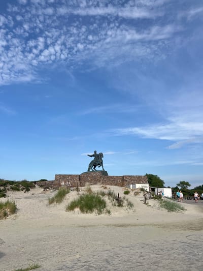The image captures an outdoor scene on a bright, partly cloudy day. In the foreground, a sandy embankment with sparse green vegetation rises towards a large stone structure. Perched atop this structure is a dark bronze equestrian statue of a man in historical attire, his right arm extended forward. The statue is set against a backdrop of a blue sky dotted with scattered white clouds.

Behind the statue, a low, rough-hewn stone wall forms the base of the monument. To the right of the statue, a paved walkway leads away from the sandy area. Several individuals are visible on this walkway, some walking and others standing near small white structures, one of which appears to have text on it. The environment suggests a coastal or elevated park setting. This monument is the Peter the Great Monument located in Baltiysk, Russia, marking the westernmost point of the country.