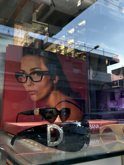 This image captures a retail display window, likely for an eyewear store, taken at an oblique angle and rotated 90 degrees clockwise. The scene combines the items inside the display with strong reflections from the exterior.
Within the display, a large poster dominates the left and center, featuring a close-up of a woman's face, shown from the nose upwards. She is looking to the viewer's left with a calm expression and is wearing a pair of modern black-framed eyeglasses. The poster has a vibrant red background. To the right of the poster, two pairs of black sunglasses are prominently displayed. One pair has a distinct angular or geometric shape with a small, gold-colored emblem on the temple. The second pair, positioned further right, features a large, decorative silver 'D' embellished with rhinestones or crystals on its temple. Partially visible behind these items are out-of-focus display stands and possibly purple packaging. The text "BANA" and "GABBANA" (reversed) is visible on the far right, suggesting a Dolce & Gabbana product or branding.
The glass of the display heavily reflects a bright outdoor urban environment, indicative of daytime with sunny weather. Reflections include a multi-story building with numerous windows and metal railings, possibly balconies or an external structure. A street lamp is also clearly reflected, along with patches of blue sky. In the upper left reflection, the text "CHANGE COLLECTIONS" is visible. The overall impression is a vibrant street scene blending with the retail presentation, located in an urban setting such as Koridallos, Greece. No direct human activity is depicted within the display or clear in the reflections.