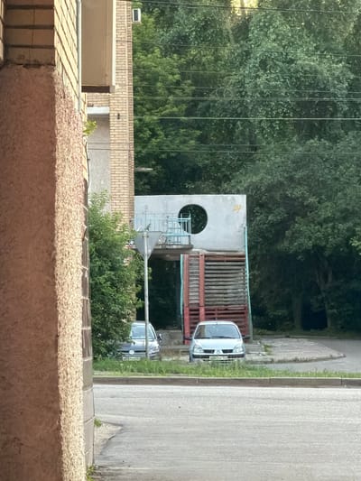 The image shows an outdoor scene, likely in Kolomna, Russia, featuring a playground structure and two cars parked on the side of a road. The playground structure is made of concrete and wood, with a circular opening in the concrete section and a wooden staircase. There are trees and foliage in the background, suggesting a natural or park-like setting. One car, a silver minivan, is parked in front of the playground, and another, a silver hatchback, is parked behind it. The scene appears to be set during daylight hours, with soft lighting that could indicate early morning or late afternoon. The overall impression is one of a quiet, residential area. There is some faint yellow spray paint on the concrete part of the playground structure. There are no people visible in the image.
