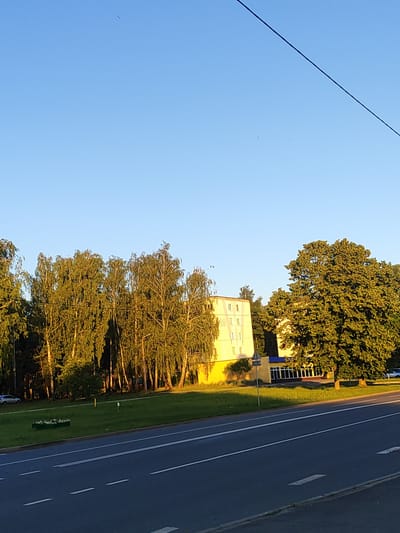 This media file captures an outdoor scene, likely in Svietlahorsk, Belarus, during the golden hour of a clear day. The sky is a vibrant, cloudless blue, with a single dark line, possibly a power cable, diagonally traversing the upper right portion of the frame. Below the sky, a dense line of tall, slender trees with green foliage forms the background. The trees appear to be illuminated by the setting sun, casting long shadows.

In the mid-ground, a bright yellow building with multiple windows is visible. To its right, a large, mature tree with a full canopy of green leaves stands prominently. Adjacent to the yellow building, a smaller structure with a blue-trimmed entrance is also present.

In the foreground, a paved road with clear white lane markings stretches across the bottom of the image. A strip of green grass separates the road from the buildings and trees. On this grassy area, a small, rectangular planter box filled with white flowers is visible on the left. A light-colored car is parked on the far left, on the other side of the grassy strip.

The overall impression is one of a peaceful, late afternoon or early evening setting in a semi-urban or suburban environment, characterized by natural light and a clear sky. There is no discernible activity taking place, and no people are visible. The scene evokes a sense of calm and tranquility.