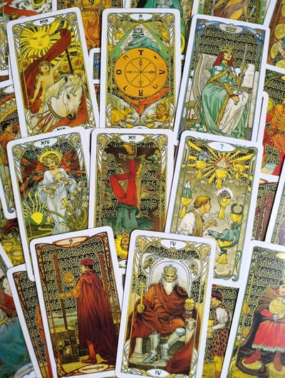 The image is a top-down, overhead view of a scattered collection of tarot cards. The cards are artfully illustrated with intricate, gilded borders and detailed depictions of figures and symbolic imagery in a style reminiscent of Art Nouveau. Several cards are clearly visible, including The Sun (XIX), The Wheel of Fortune (X), The Empress (III), The Hanged Man (XII), Temperance (XIV), the Seven of Cups (VII), the Two of Wands (2), the King of Pentacles (IV), and others partially obscured. The cards are arranged on a flat surface, overlapping each other, with no discernible background beyond the edges of the cards themselves. There is no information in the image to indicate the city of Alexandrov, Russia, or any other specific location. The scene is static and appears to be a still life arrangement of tarot cards.